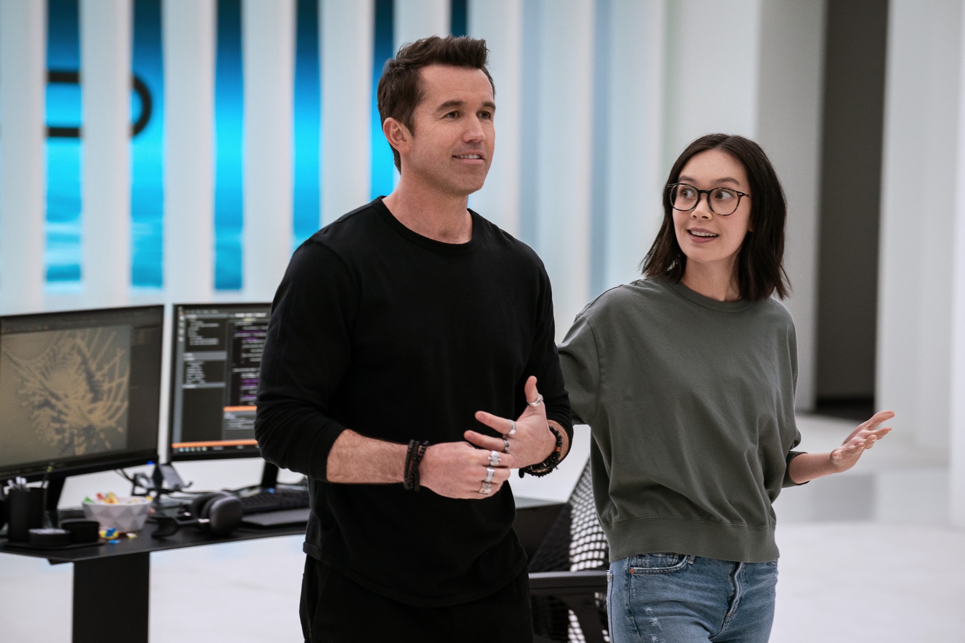 Rob McElhenney y Charlotte Nicdao sonríen mientras caminan por una oficina en una escena de la temporada 3 de Mythic Quest.