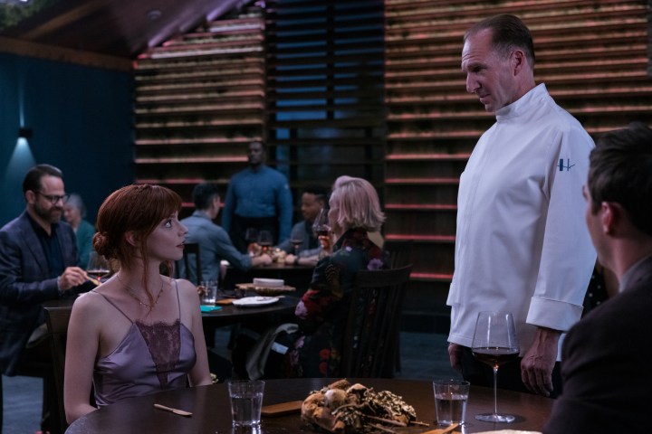 Anya Taylor-Joy sits down and looks up at Ralph Fiennes in a scene from The Menu.