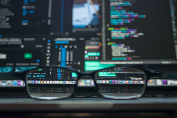 A pair of glasses rests on a desk in front of multiple computer monitors filled with code.