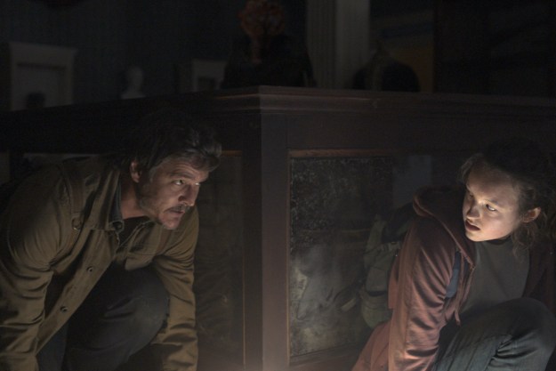 Pedro Pascal and Bella Ramsey crouch behind a glass case together in The Last of Us.