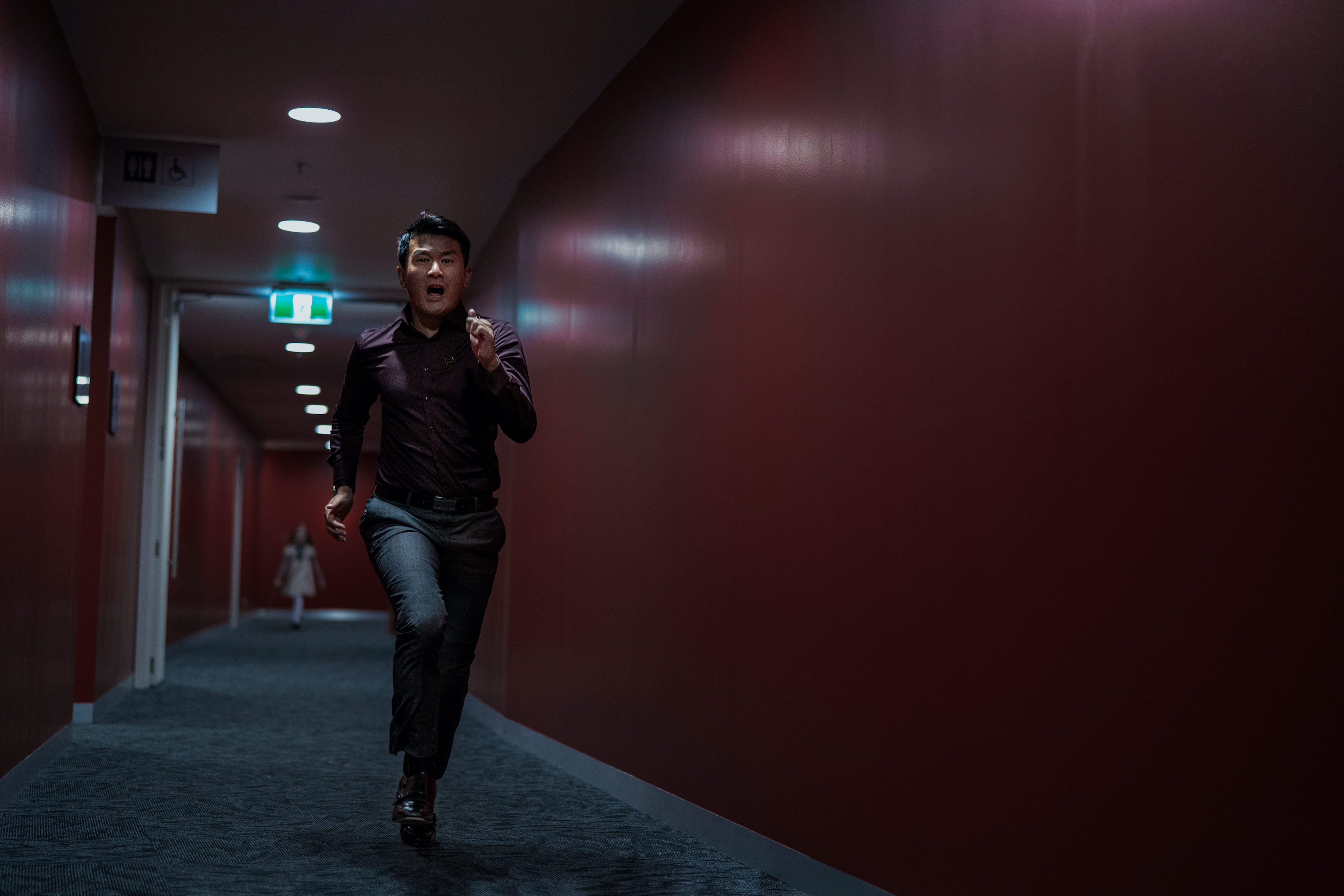 Ronny Chieng corre por un pasillo rojo alejándose de M3GAN.