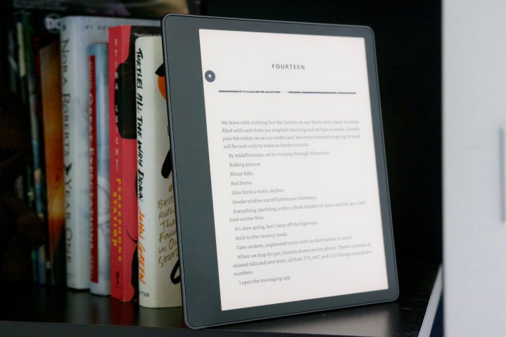 Amazon Kindle Scribe sitting on a bookshelf next to a stack of books.