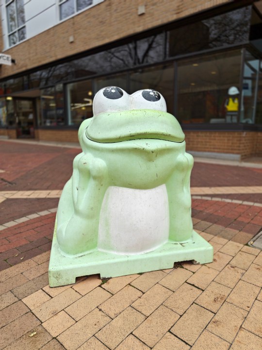 Portrait mode photo of a frog sculpture on a brick walkway.