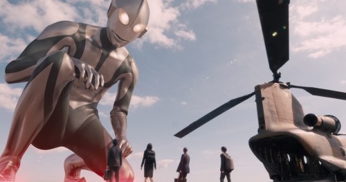 A giant being, Ultraman, bends down to look at four humans on the ground next to a helicopter.