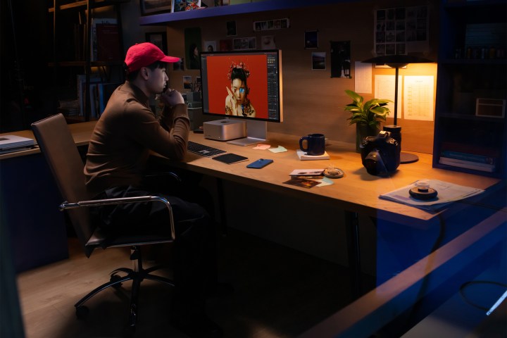 Uma pessoa sentada em uma mesa usando um Apple Mac Studio ao lado de um Studio Display.  Há um baixo nível de iluminação com vários objetos colocados sobre a mesa.