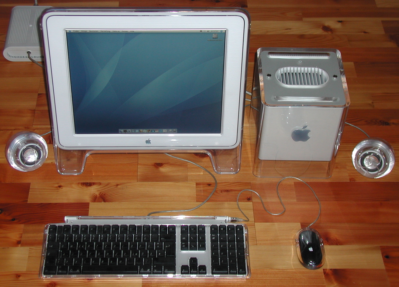An Apple Power Mac G4 Cube computer on a desk next to an Apple monitor, keyboard, mouse and speakers.