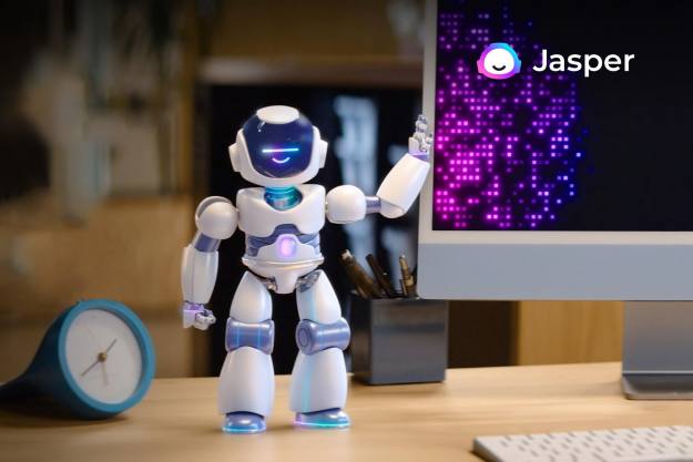 Jasper AI's robot mascot stands on a desk by a computer.