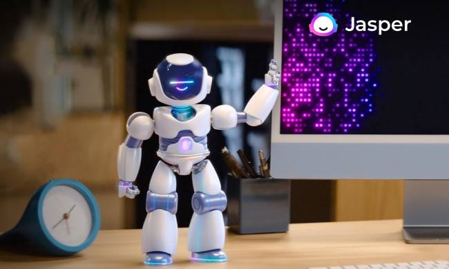 Jasper AI's robot mascot stands on a desk by a computer.
