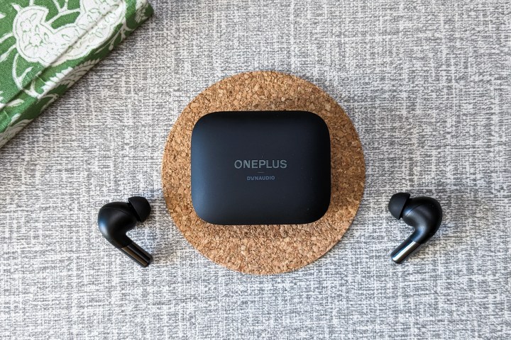 Black OnePlus Buds Pro 2 case on a cork coaster and black headphones on a white and gray table top.