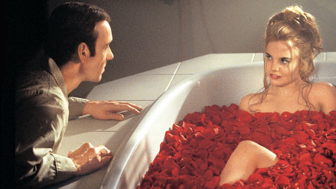 Kevin Spacey agachado ao lado de uma jovem em uma banheira, coberto de pétalas de rosa em uma cena de American Beauty.