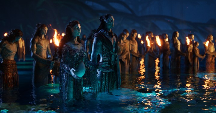A group of Na'vi mourn in the water while flames show around them in a scene from Avatar: The Way of Water.