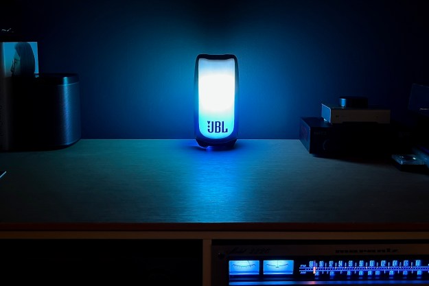 The JBL Pulse 5 Bluetooth speaker, glowing blue on a media sideboard table with a stereo receiver.