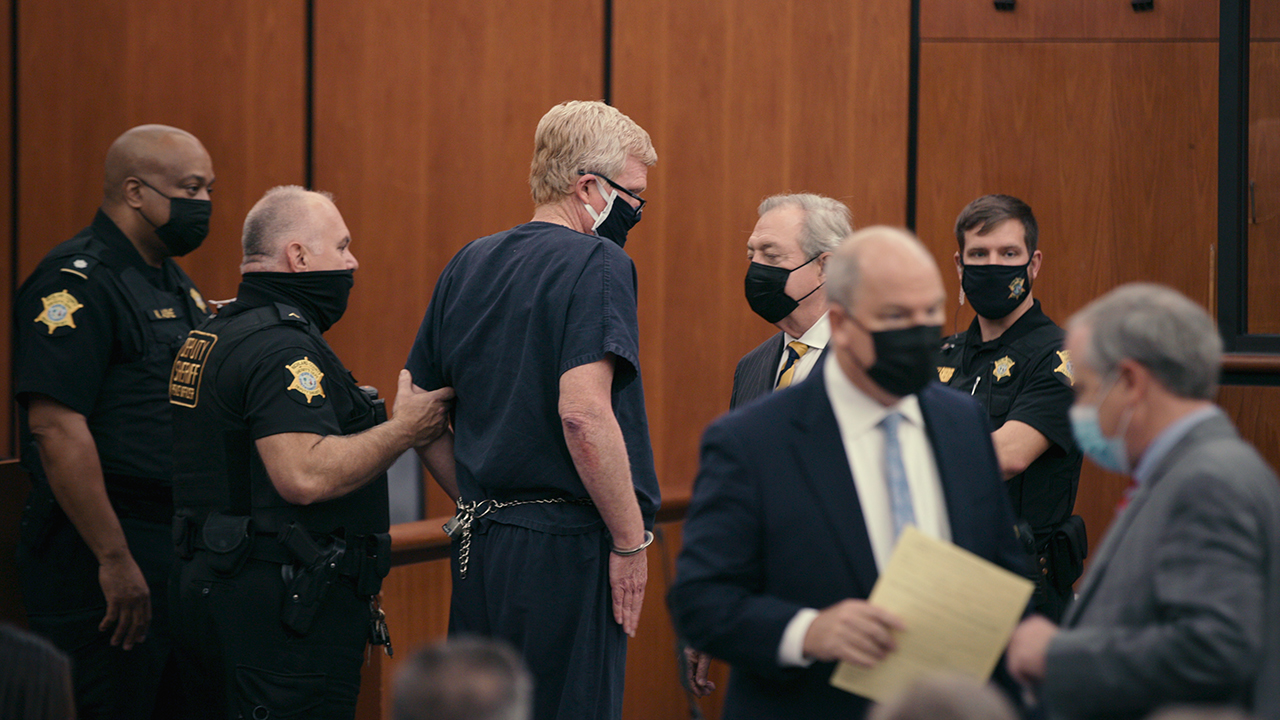 Alex no tribunal vestindo um macacão de prisão e máscara com outras pessoas ao seu redor em uma cena de Murdaugh Murders na Netflix.