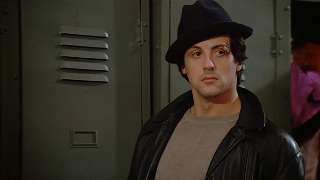 Rocky Balboa parado ao lado de um armário com um chapéu, o olho machucado em uma cena de Rocky.