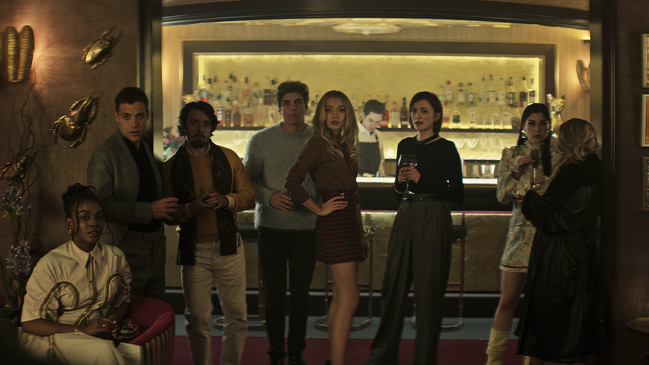 Os novos personagens da 4ª temporada de You parados em um bar, vestidos com esmero, olhando para alguma coisa.