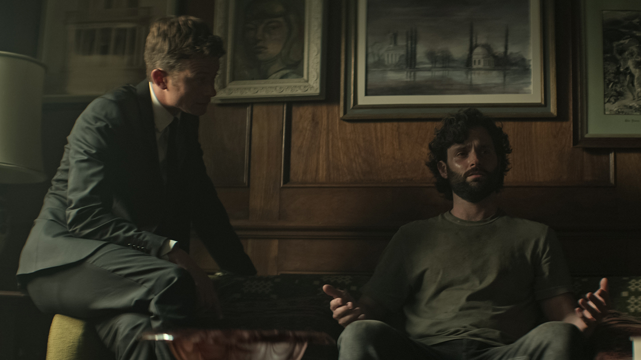 Joe perturbado no sofá, Rhys ajoelhado sobre ele em uma cena da 4ª temporada de You.