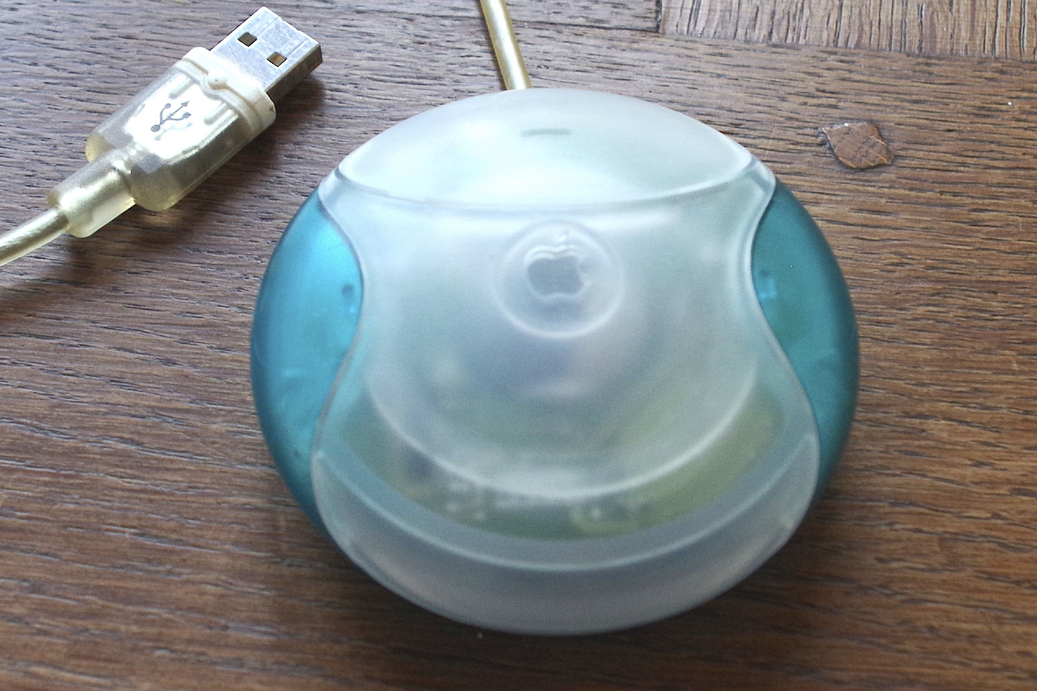 Um mouse USB da Apple, conhecido como mouse "disco de hóquei", do iMac G3.  O mouse está sobre uma mesa com o cabo USB ao lado.