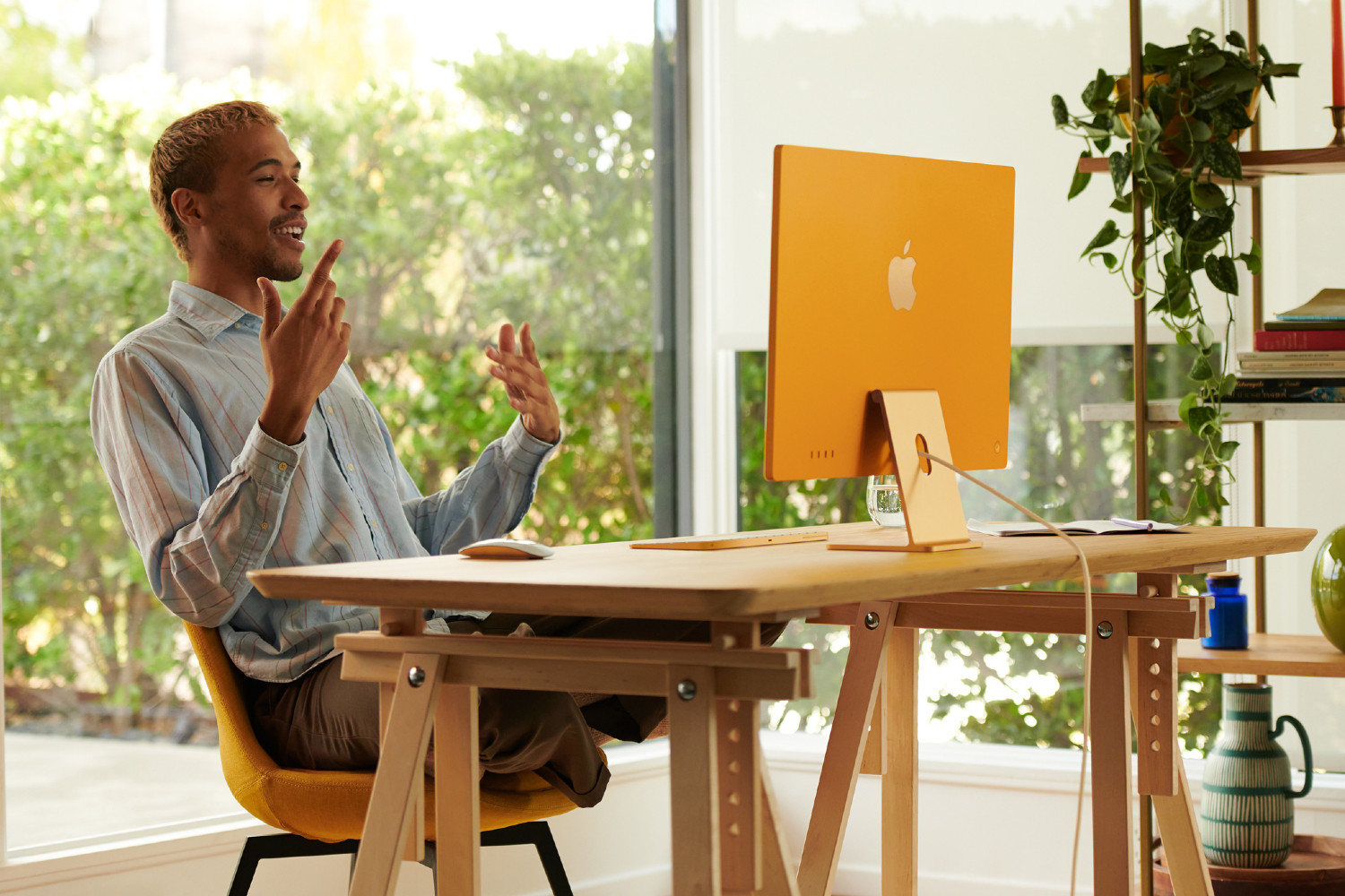 一名男子坐在 M1 iMac 前的办公桌前。他身后是一扇大玻璃窗和一组架子，架子上放着书籍、植物和装饰品。