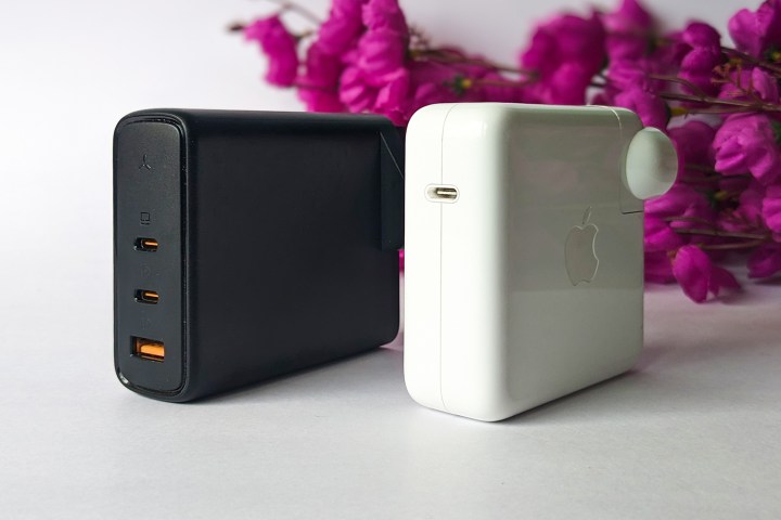 Voltme Revo 140 GaN charger beside Apple's 61W MacBook Pro charger in front of pink fake flowers against a white background. 