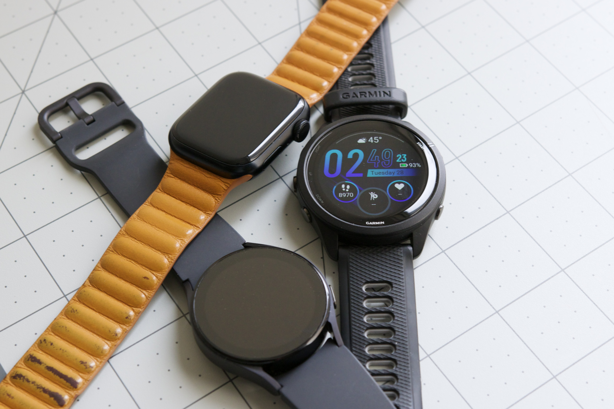 Apple Watch، Galaxy Watch 5 و Garmin Forerunner 265 کنار هم روی یک میز قرار دارند.