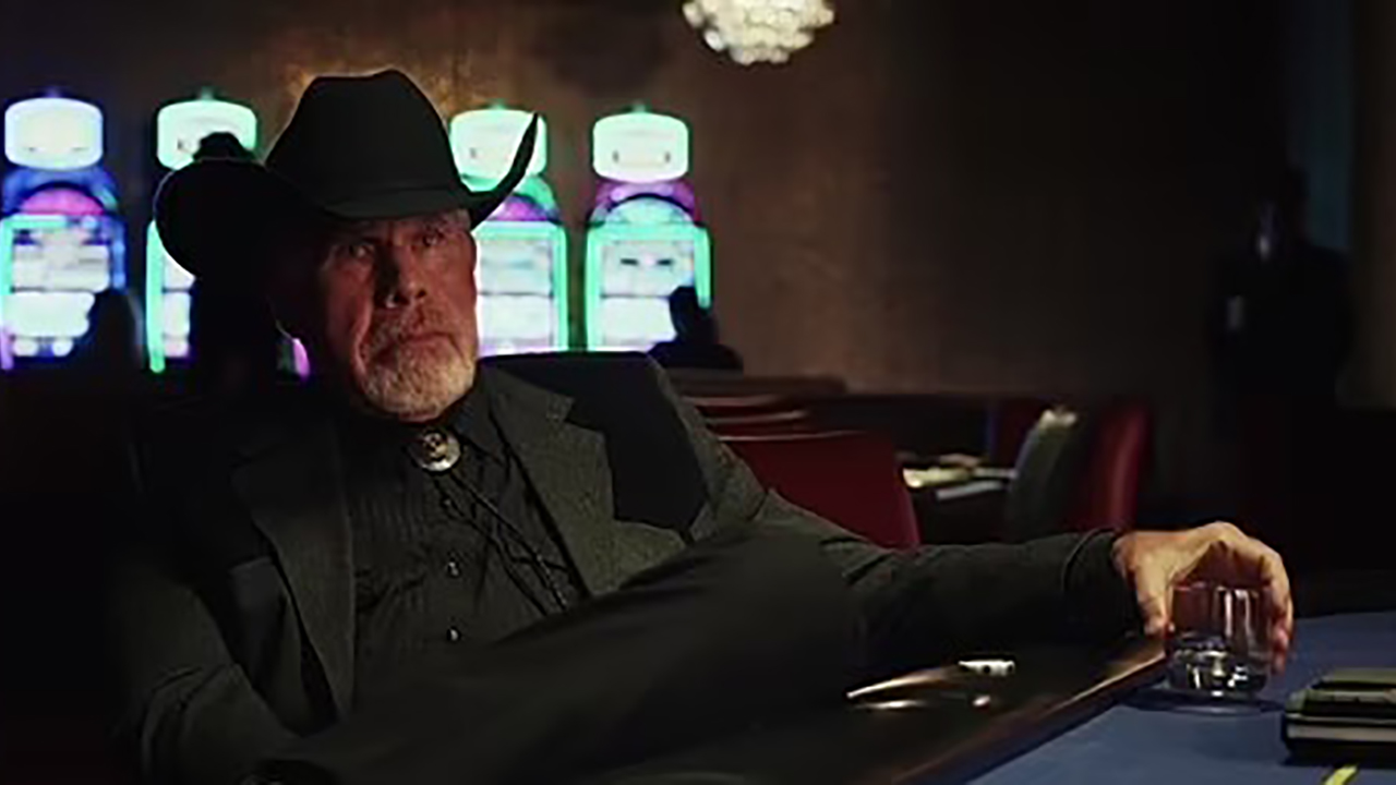 Ron Perlman com um chapéu de cowboy sentado em uma mesa de pôquer segurando uma bebida em uma cena de Poker Face on Peacock.