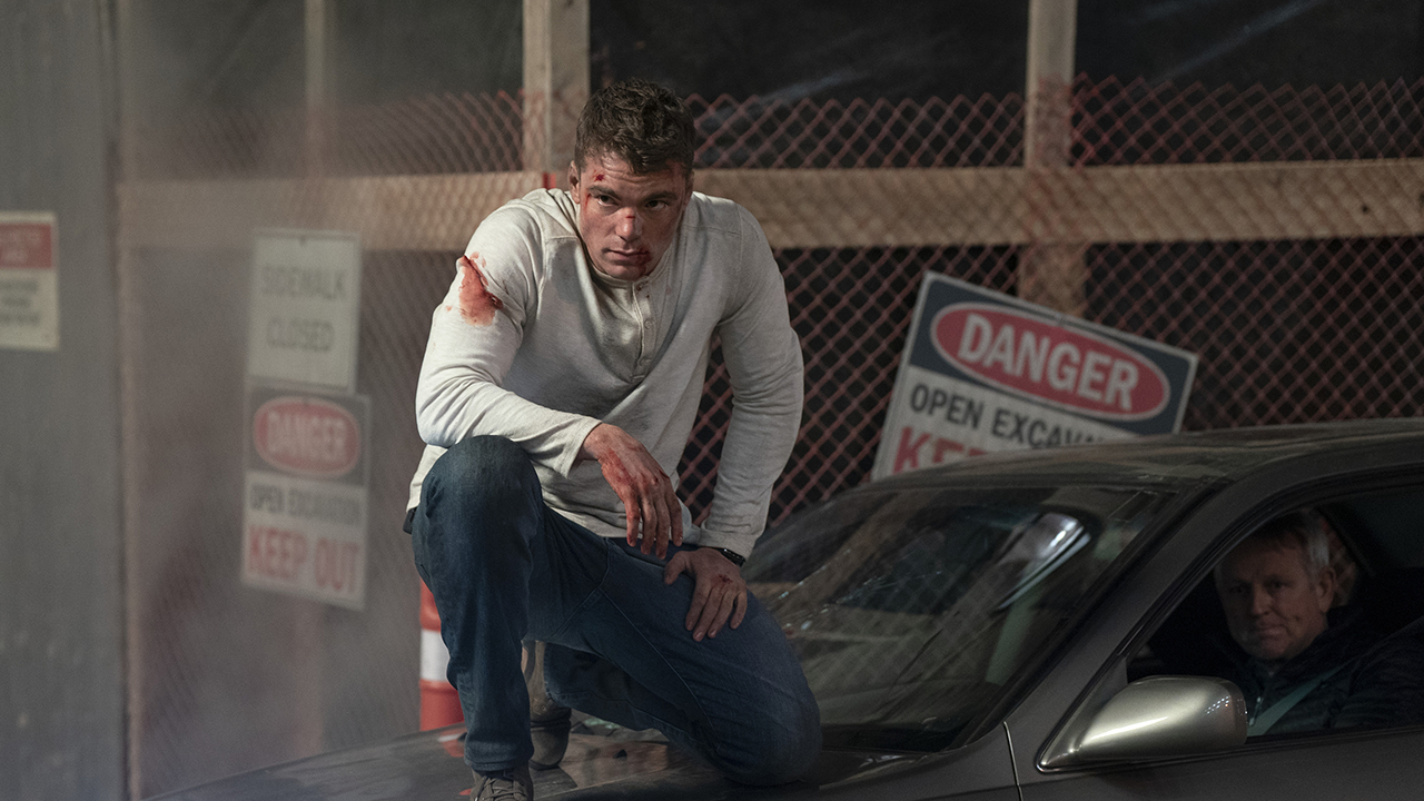 Peter ajoelhado em cima de um carro, ensanguentado e espancado em uma cena de The Night Agent.