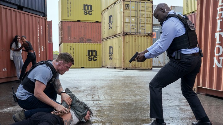 Peter sujetando a Colin, el agente Monks apuntando con un arma a la suya, Maddie y Chelsea en el fondo de un astillero en una escena de The Night Agent.