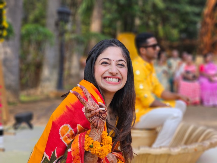 Indian bride smiling and making a victory sign with her hand with groom in the background captured with the Samsung Galaxy S23 Ultra’s 3x telephoto portrait mode camera.