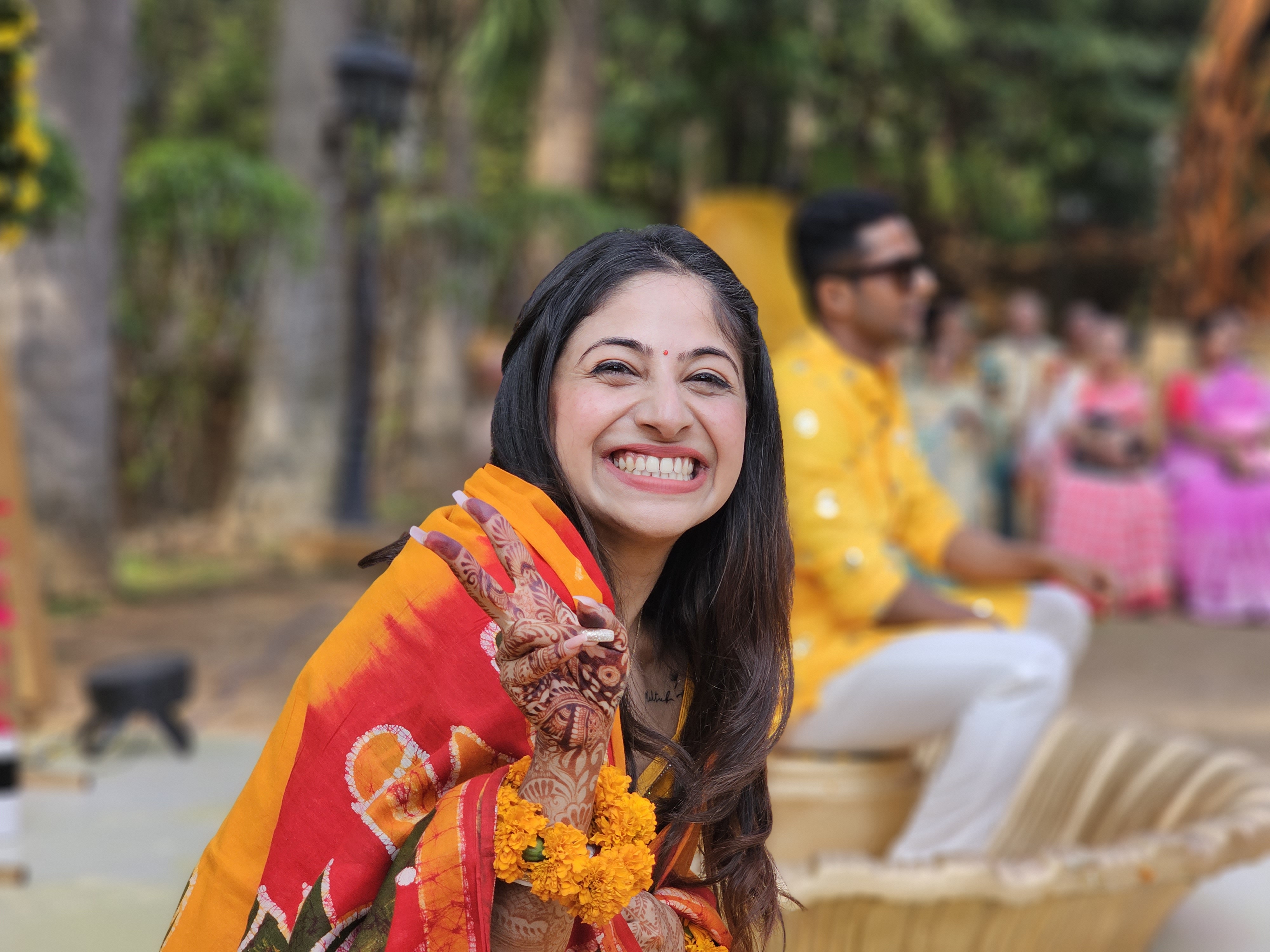 Noiva indiana sorrindo e fazendo um sinal de vitória com a mão com o noivo ao fundo, capturado com a câmera telefoto 3x no modo retrato do Samsung Galaxy S23 Ultra.