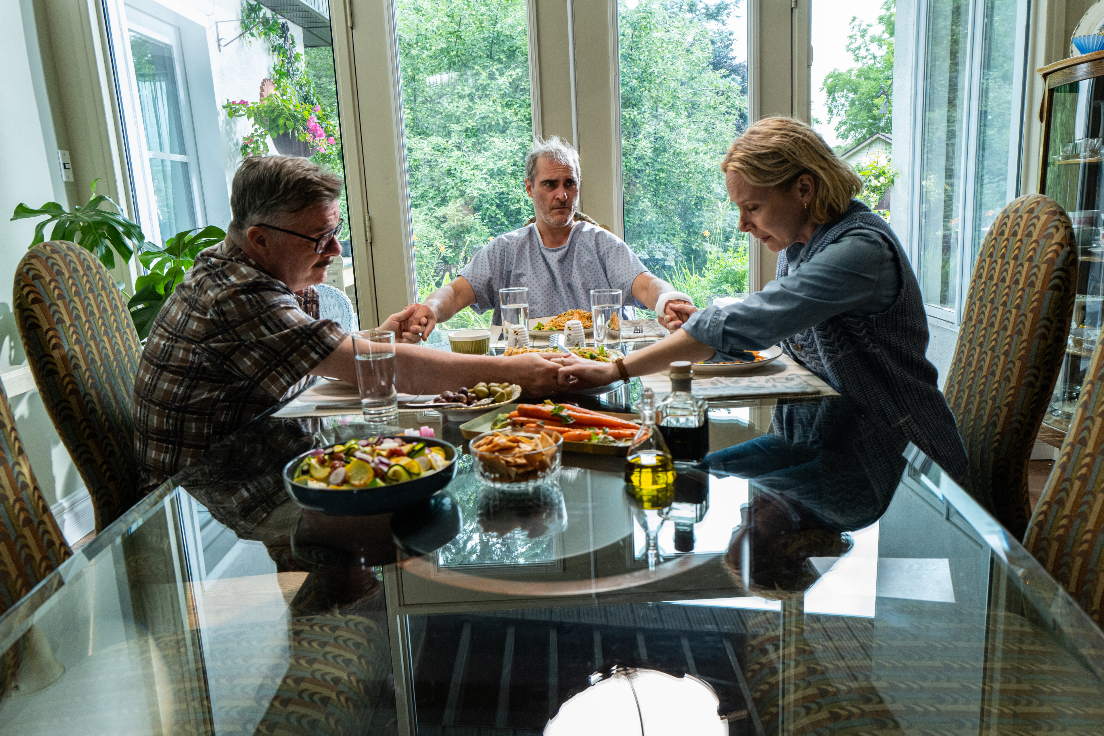 Nathan Lane, Joaquin Phoenix e Amy Ryan sentam-se ao redor de uma mesa juntos em Beau is Afraid.