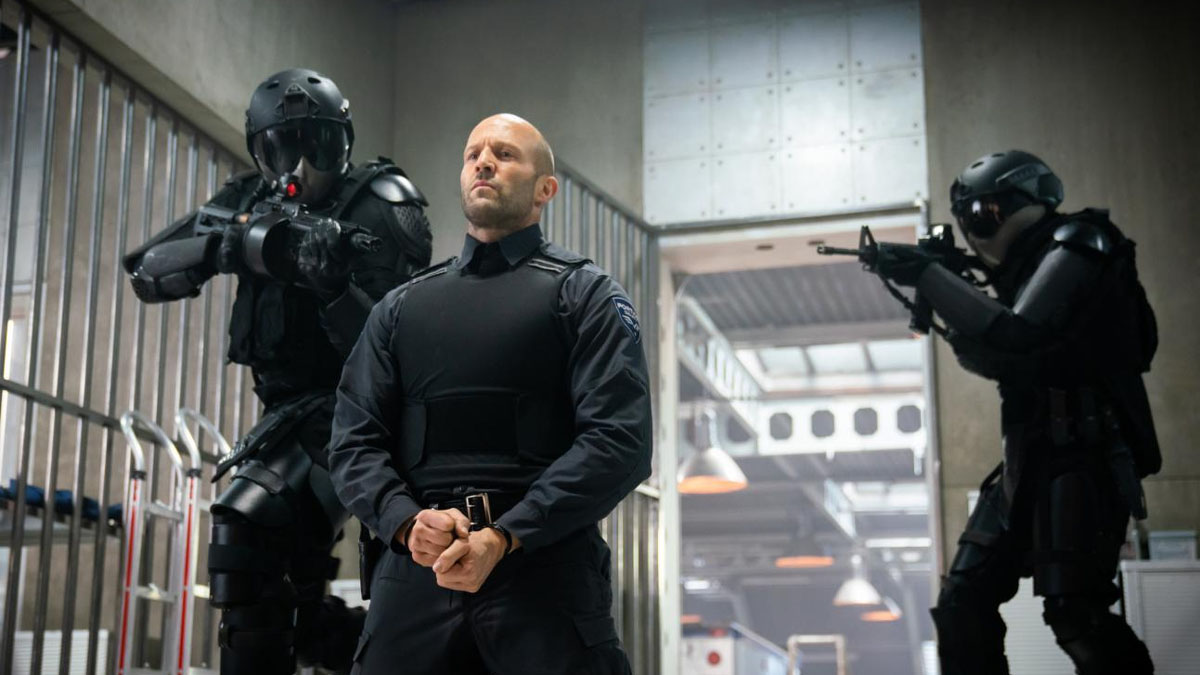 Jason Statham está sentado, algemado, na frente de dois guardas em Wrath of Man.