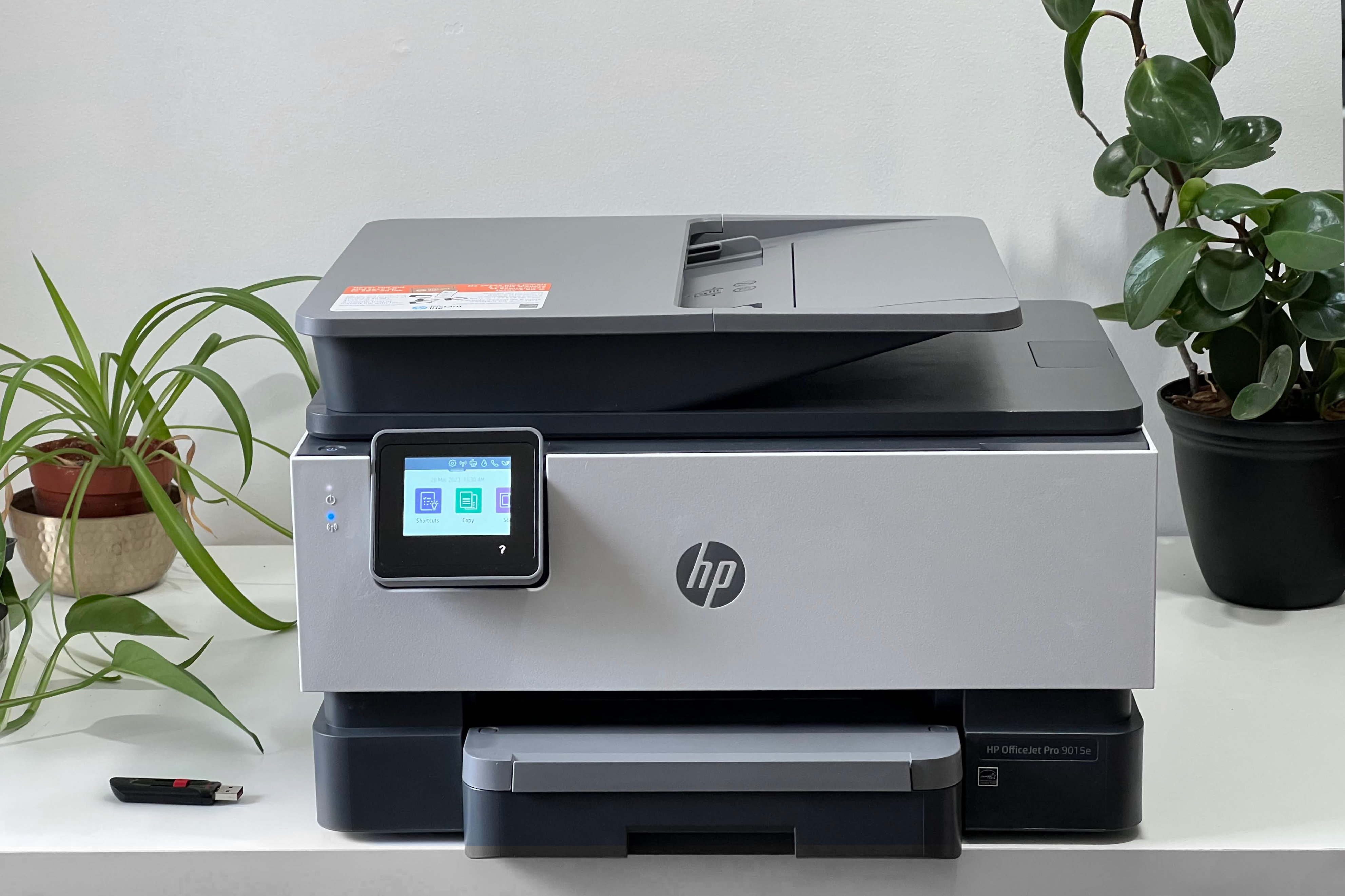 Une imprimante tout-en-un HP OfficeJet Pro 9015e repose sur une table blanche avec des plantes et une clé USB à côté.