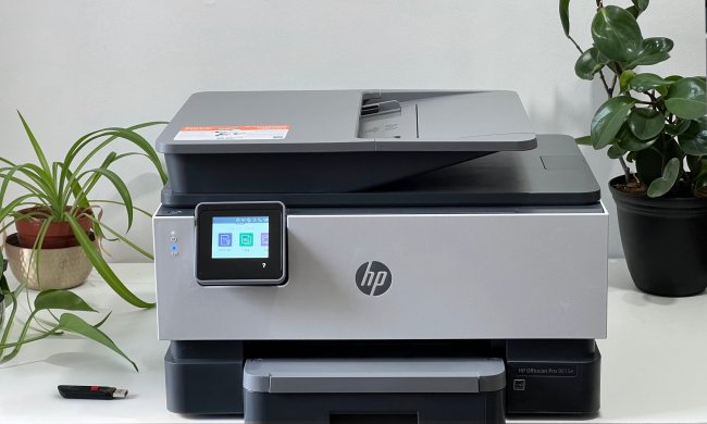 An HP OfficeJet Pro 9015e all-in-one printer rests on a white table with plants and a thumb drive beside it.