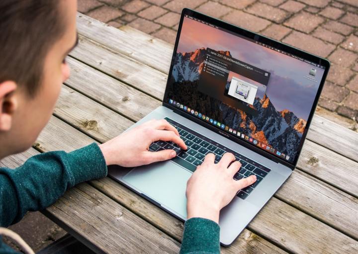 A person typing on a MacBook Pro while sat on a wooden bench.