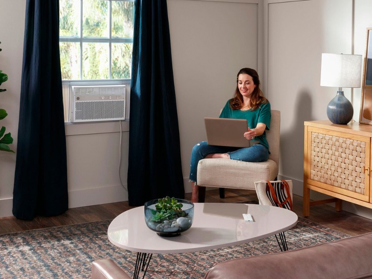 Uma mulher trabalha em um laptop enquanto um ar-condicionado de janela inteligente GE 8.000 BTU resfria o ambiente.