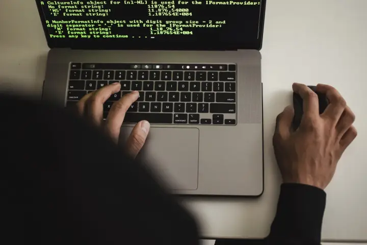 A hacker typing on an Apple MacBook laptop, which shows code on its screen.