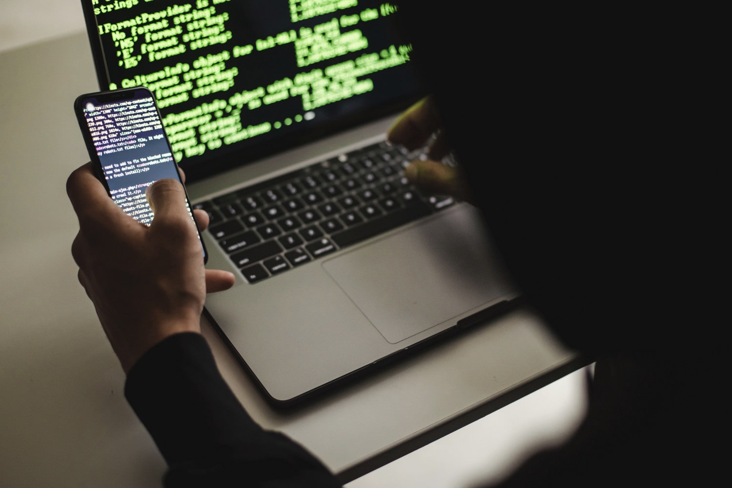Un hacker escribiendo en una computadora portátil Apple MacBook mientras sostiene un teléfono. Ambos dispositivos muestran código en sus pantallas.