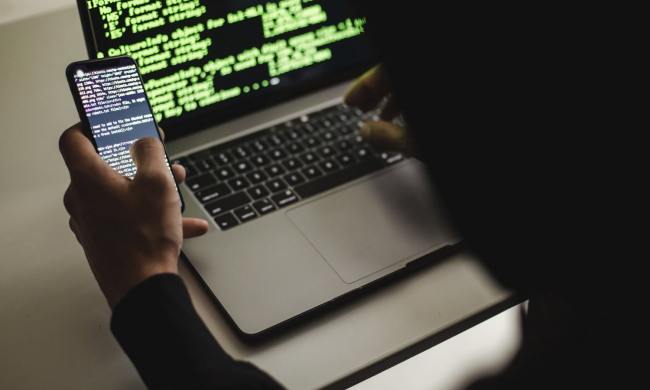 A hacker typing on an Apple MacBook laptop while holding a phone. Both devices show code on their screens.