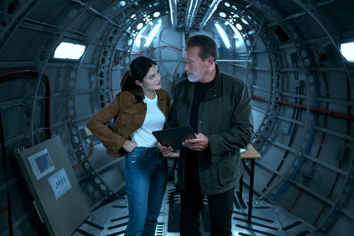 Monica Barbaro and Arnold Schwarzenegger stand on a cargo plane together in FUBAR.