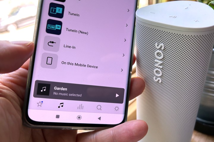 An Android phone with the Sonos app showing the music sources tab menu, next to a Sonos Roam wireless speaker.