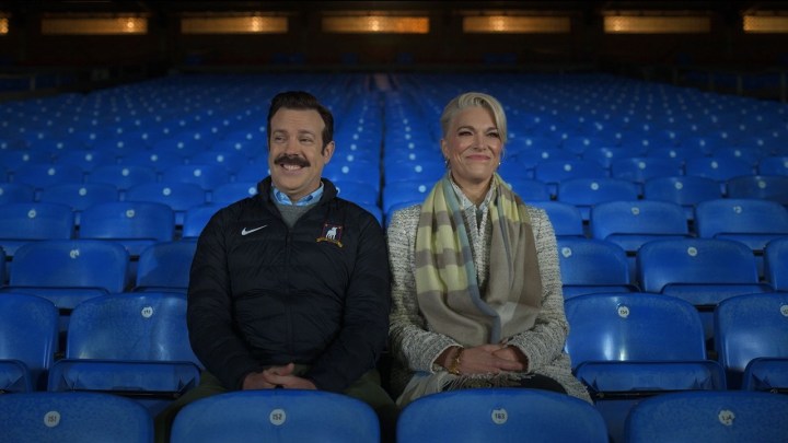 Ted and Rebecca sitting in stadium seats laughing together in a scene from Ted Lasso.