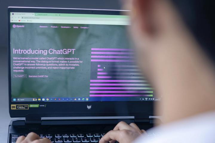 Человек сидит перед портативным компьютером. На экране ноутбука находится домашняя страница чат-бота OpenAI ChatGPT с искусственным интеллектом.