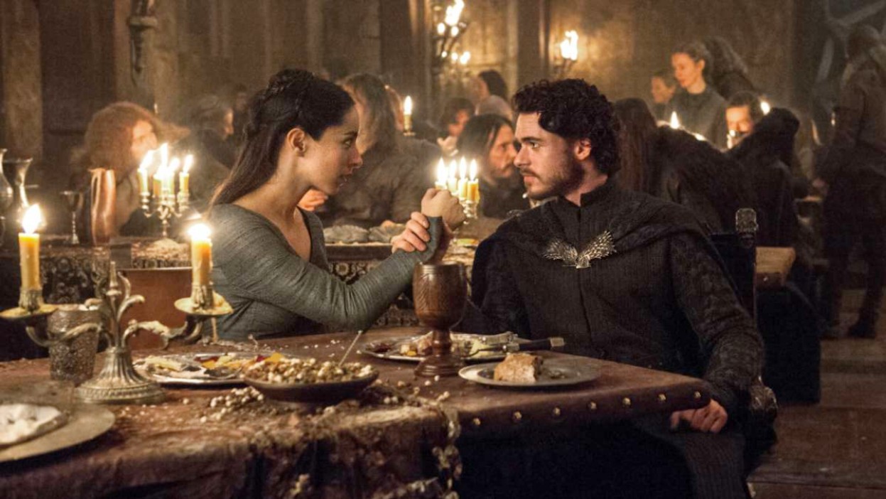 Oona Chaplin and Richard Madden as Talisa and Robb Stark holding hands at a dinner party in Game of Thrones.