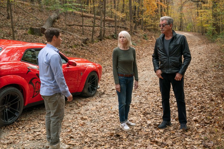 Adam Devine, Ellen Barkin, and Pierce Brosnan stand near a red muscle car in The Out-Laws.