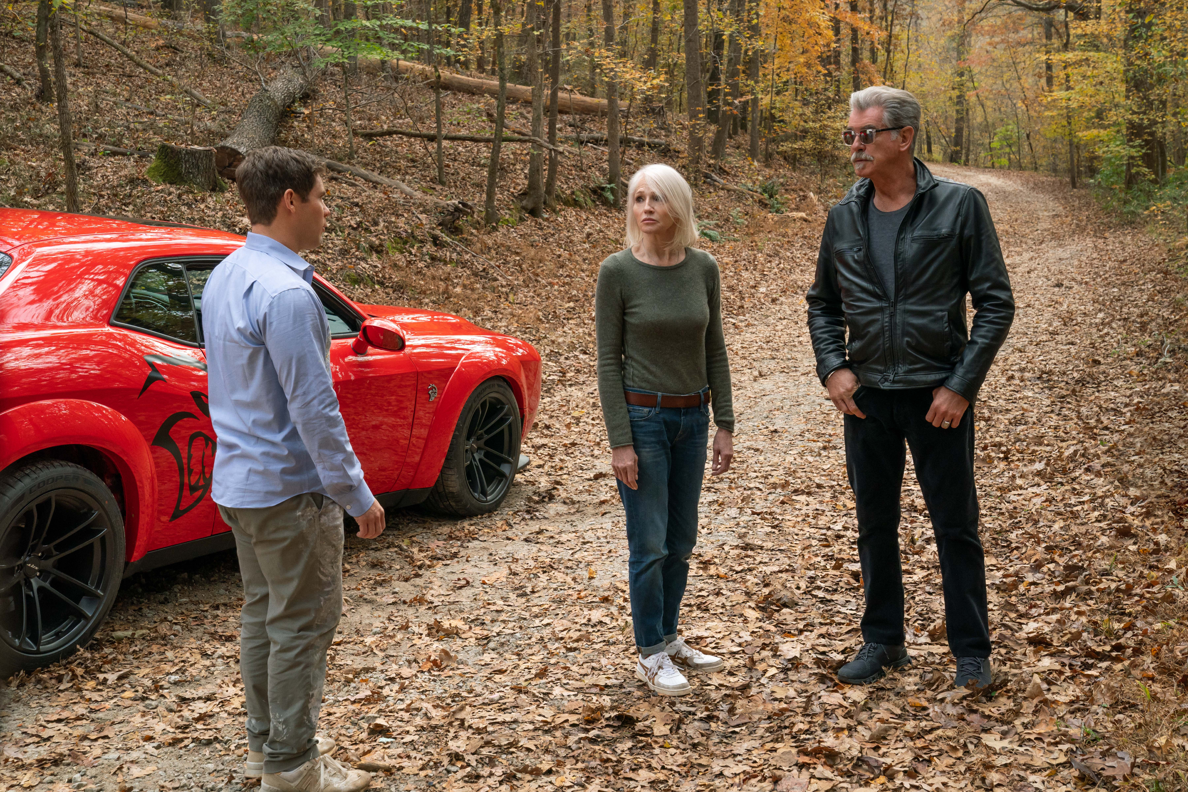 Adam Devine, Ellen Barkin e Pierce Brosnan estão perto de um muscle car vermelho em The Out-Laws.
