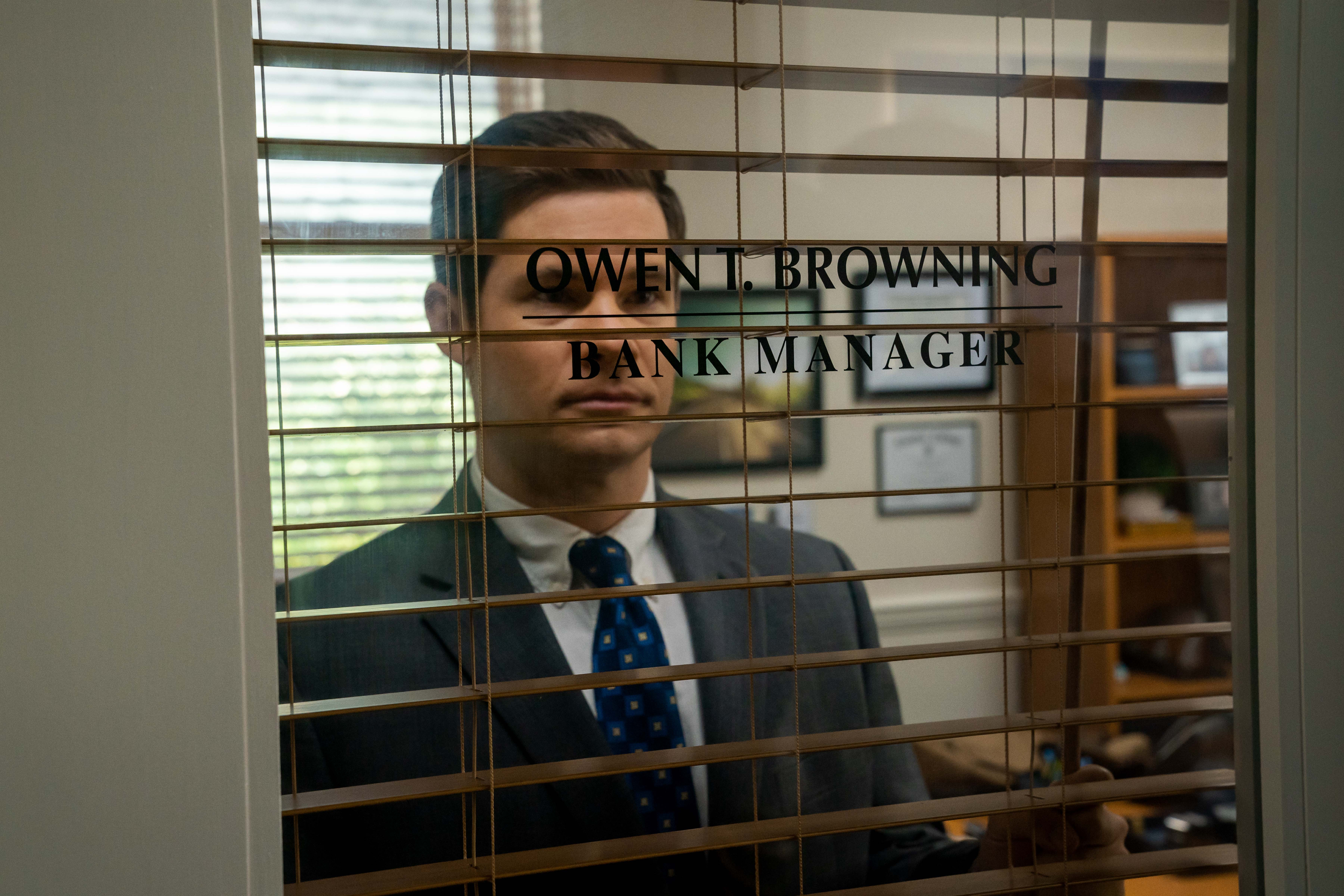 Adam Devine olha pela janela de seu escritório no banco em The Out-Laws.
