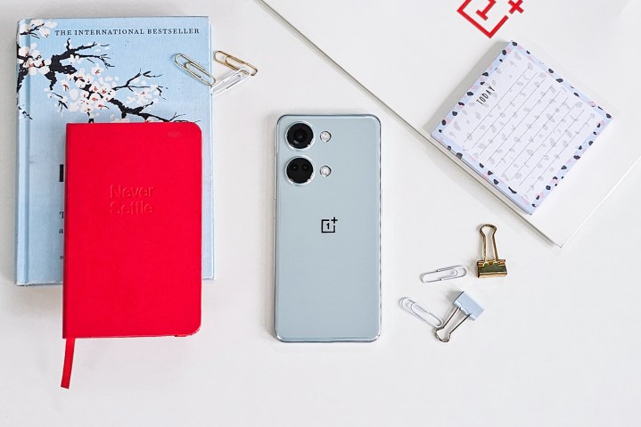 OnePlus Nord Misty Green color on white background with a red Never Settle notebook, post it notes, clips, and a blue book on the side.