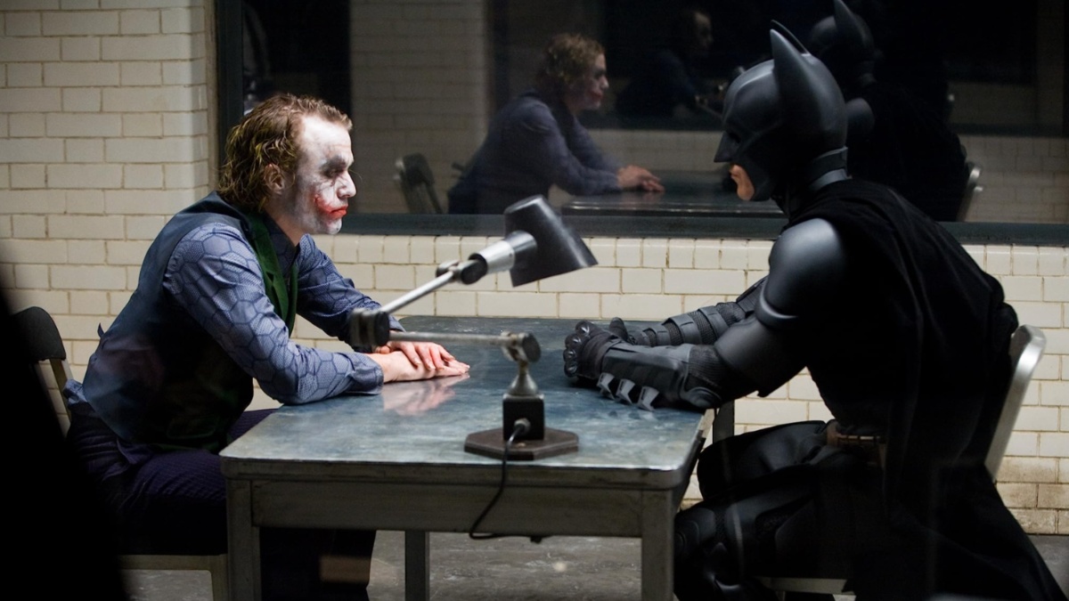 O Coringa e o Batman sentam-se frente a frente em uma mesa.