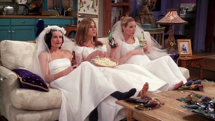 Monica, Rachel, and Phoebe sitting on the couch in wedding dresses drinking beer and eating popcorn in a scene from Friends.