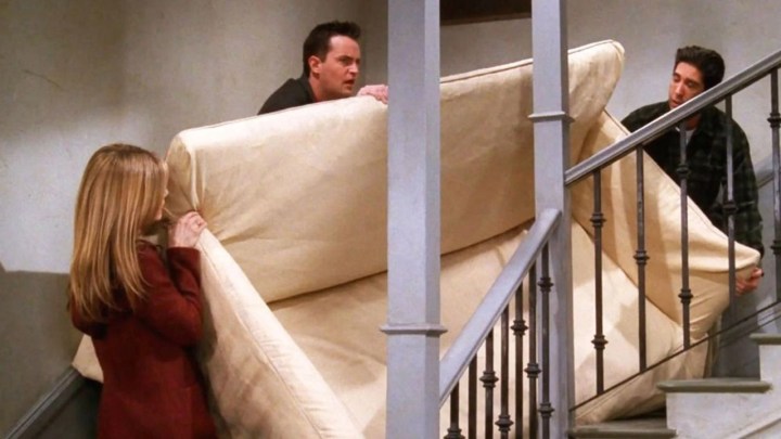 Rachel, Chandler, and Ross standing at different sides of a couch, trying to move it up a set of stairs on Friends.